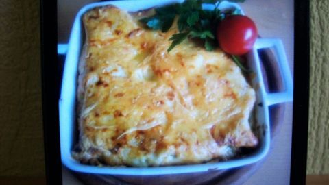 Eine goldbraune Portion gratinierte Spinat-Lachs-Pfanne liegt in einer weißen Auflaufform auf einem Holzbrett, garniert mit Petersilie und einer Kirschtomate.