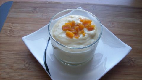 Ein Glas Mango-Joghurt mit Aprikosenstücken steht auf einem weißen Teller, umgeben von einer Holzplatte.
