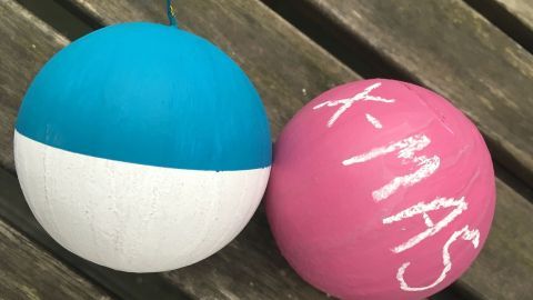 Bunte Weihnachtskugeln mit Tafellack, eine in Blau und Weiß, die andere in Pink mit Kreidebeschriftung "X-MAS", liegen auf einer Holzoberfläche.