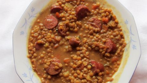 Ein Teller deftiger Linseneintopf mit Linsen und Würstchen liegt appetitlich angerichtet auf einem weißen Tisch.