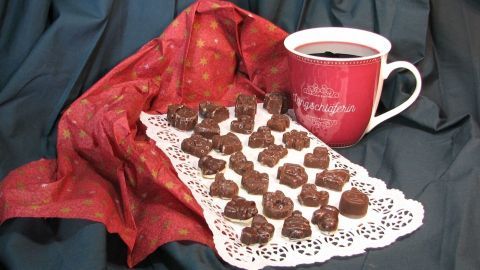 Schokoladenpralinen liegen auf einer Serviette, während eine Tasse Eisschokolade im Hintergrund steht, umgeben von rotem Stoff.