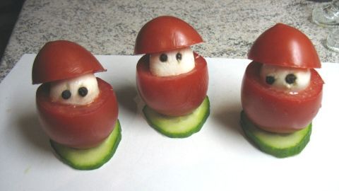 Drei Tomaten-Mozzarella-Weihnachtsmänner stehen auf einem weißen Teller, umgeben von Gurkenscheiben.