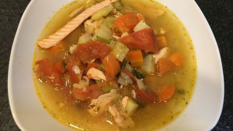 Ein Teller Lachssuppe mit buntem Gemüse wird serviert, in einer weißen Schüssel auf einem dunklen Tisch.