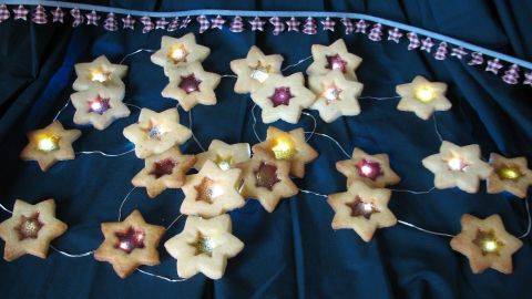 Leckere Mürbteiggebäcke in Sternform liegen dekorativ auf einem dunklen Tuch, umgeben von lichterfüllten Weihnachtsmotiven.
