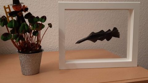 Ein schwebender Rahmen zeigt eine schwarze Skulptur, während ein kleines Pflänzchen daneben in einem Topf steht.