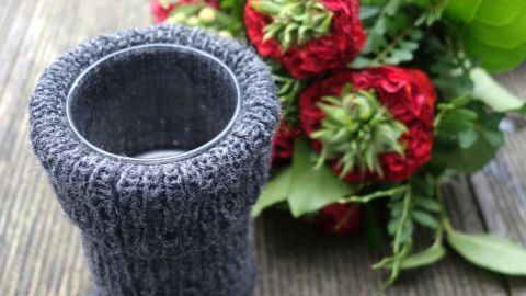 Ein grauer Vasenüberzug steht neben einem bunten Blumenstrauß auf einem Holztisch, umgeben von frischem Grün.
