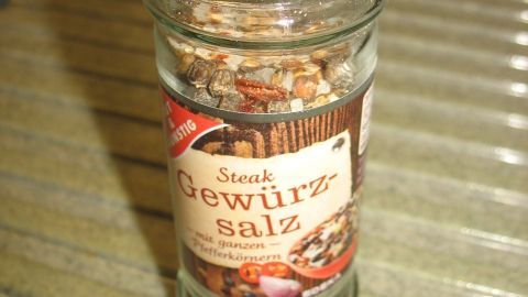 Ein Glasgewürzstreuer mit der Aufschrift „Gewürzsalz“ steht auf einer glatten Oberfläche und enthält eine Mischung aus Salz und Kräutern.