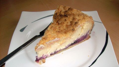 Eine große Stück Käsekuchen mit Mandelstreusel und Heidelbeeren liegt auf einem weißen Teller, bereit zum Genießen.