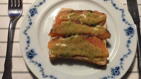 Zwei Scheiben Gravad Lax auf einem Teller, garniert mit Honig-Dill-Soße, neben Besteck auf einem gestreiften Tisch.