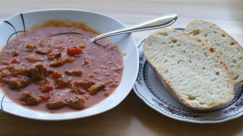 Eine dampfende Schüssel Gulaschsuppe mit Stückchen und einer Gabel steht neben zwei Scheiben frischem Brot auf einem Holztisch.
