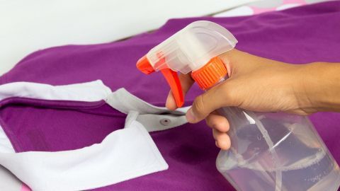 Eine Hand sprüht ein Reinigungsmittel auf ein lila Kleidungsstück, um Schweißgeruch aus der Kleidung zu entfernen.