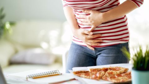 Eine Person mit Bauchschmerzen hält sich den Bauch und steht neben einer Pizzaschachtel, während sie in einem modernen Raum sitzt.