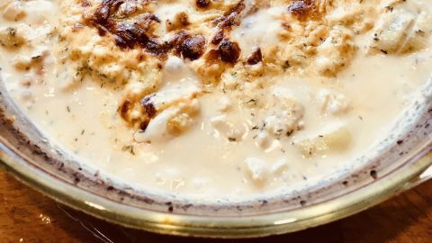 Der Lachs-Krabbengratin hat eine goldbraune Oberfläche und liegt in einer Glasform auf einem Holzbrett, umgeben von frischen Kräutern.