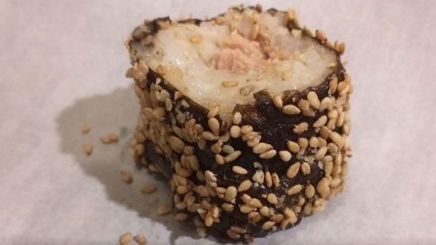 Ein Stück Sushi liegt auf einem weißen Papier. Es besteht aus Reis und ist mit Sesam umhüllt, wirkt frisch zubereitet.