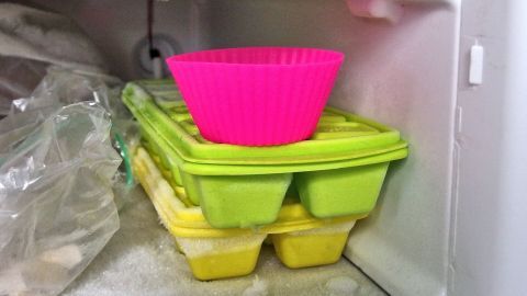 Eine pinke Muffinform steht auf grünen und gelben Eiswürfelschalen im Gefrierfach, bereit für das einfache Lösen von Muffins.