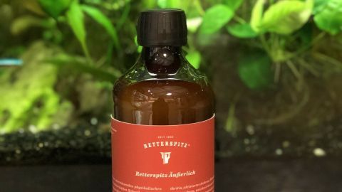 Eine braune Flasche mit einem roten Etikett steht vor einem grünen Hintergrund, umgeben von Pflanzen und Wasser.