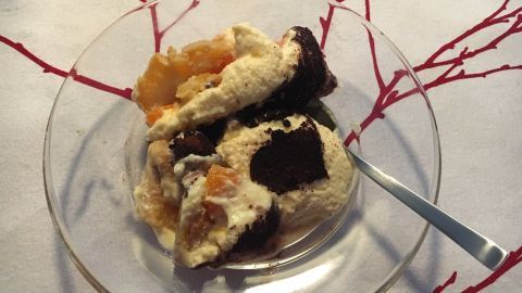 Ein Tiramisu mit Mandarinen liegt in einer Glas-Schüssel und ist mit Schokoladensauce garniert, umgeben von einem weißen Tisch und roten Ästen.