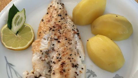 Ein gebratener, fettarmer Fisch liegt auf einem Teller, neben gekochten Kartoffeln und Limettenscheiben.
