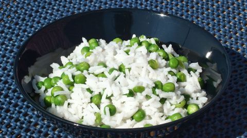 Ich gebe gerne Erbsen zum Reis und koche alles zusammen. So werden die Vitamine der Erbsen vom Reis aufgenommen und werden nicht mit dem Kochwasser weggeschüttet. Eine Schüssel Reis mit Erbsen steht auf einer blauen Unterlage, während die grüne Erbsenfüllung den weißen Reis ergänzt.