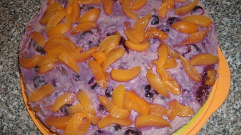 Ein saftiger Obstkuchen mit einer lila Füllung, dekoriert mit orangefarbenen Fruchtstücken, steht auf einer grauen Arbeitsfläche.