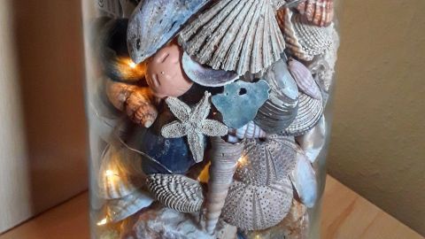 Ein Glasbehälter zeigt eine dekorative Anordnung von Muscheln und Sand, die an Urlaubserinnerungen erinnert.