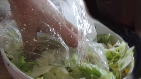 Eine Hand greift durch eine durchsichtige Folie in eine Schüssel mit frischem Salat, um ihn zu mischen.