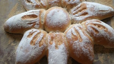Ein Brot in Form einer Sonnenblume liegt auf einer Holzfläche, leicht mit Mehl bestäubt und sorgfältig geformt.