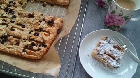 Ein frisch gebackener Apfelkuchen mit Joghurt und Mandelmilch liegt auf einem Teller, umgeben von weiteren Kuchenstücken und einer Tasse Kaffee.