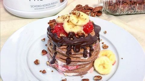 Pancakes liegen auf einem Teller und werden mit Schokolade, Banane und Nüssen garniert, umgeben von Proteinpulver.