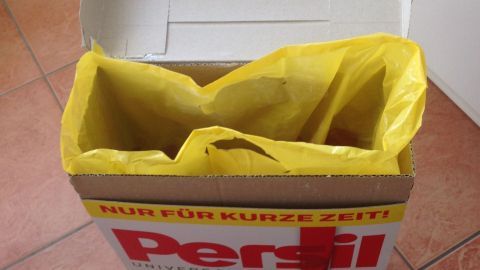 Ein offener Karton mit gelber Innenverpackung steht auf einem Fliesenboden und enthält Waschmittel, um es frisch zu halten.