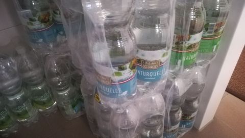 Vor einem Regal sind gestapelte Wasserflaschen zu sehen, die zur Konservierung von Trinkwasser für Notfälle bereitstehen.