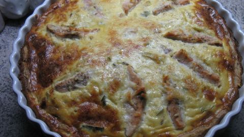 Eine goldbraun gebackene Lauch-Tarte mit Lachs liegt in einer weißen Tarte-Form auf einer grauen Arbeitsfläche.