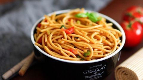 Ein schwarzer Schüsselchen Chinesischer Nudelsalat mit Paprika und Kräutern steht auf einem Tisch, daneben Tomaten.
