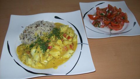 Eine Schüssel Fisch-Curry mit Wildreis auf einem Teller, daneben ein kleiner Salat, alles auf einem Tisch arrangiert.
