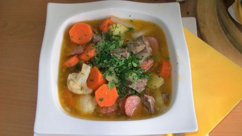 Ein bunter Gemüsetopf mit Karotten, Kartoffeln und Fleisch liegt in einer weißen Schüssel auf einem Tisch, umgeben von Besteck.