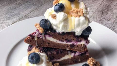Ein verführerisches Stück Schokoladen-Blaubeer-Cheesecake ist auf einem weißen Teller angerichtet, garniert mit Sahne, Blaubeeren und einer Honigglasur.