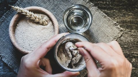 In einer handgefertigten Schüssel wird Sauerteig mit Wasser vermischt. Nebenan liegen Weizenkörner auf einem textilen Untergrund.