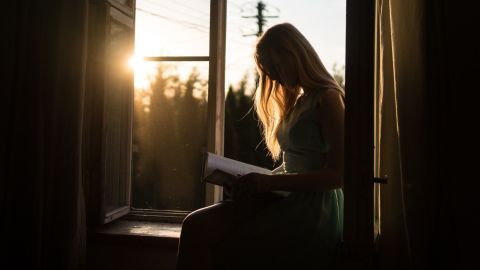 Eine Person sitzt am Fenster und liest ein Buch, während die Abendsonne durch die Öffnung scheint.