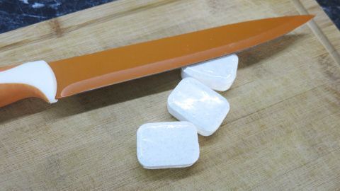 Auf einem Holzbrett liegen halbierte Geschirr-Reinigertabs neben einem Küchenmesser, bereit zum Einsatz in der Spülmaschine.