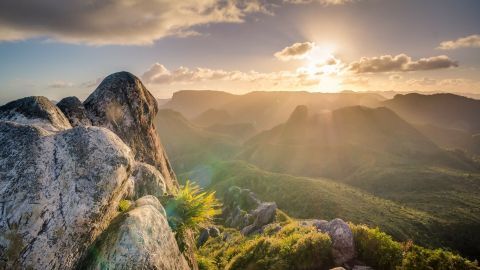 Ein majestätischer Felsen steht im Vordergrund, während die Sonne hinter den Bergen aufgeht und Hoffnung in die Landschaft bringt.
