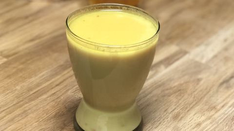 Ein Glas Goldene Milch steht auf einem Holztisch, umgeben von einer Tüte Kurkuma-Pulver und einem Glas Honig.