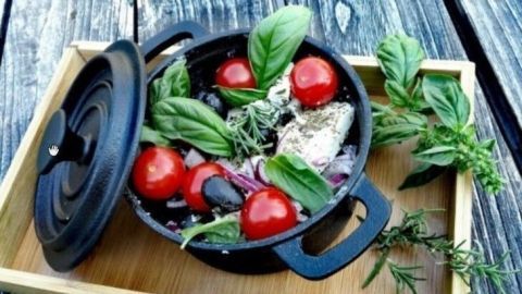 In einem schwarzen Topf liegen frische Kräuter, rote Tomaten und anderes Gemüse, bereit zum Kochen für das Fetakäse-Grillrezept.