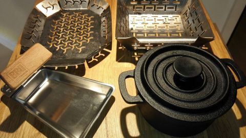 Grillzubehör liegt auf einer Holzoberfläche, einschließlich einer gitterförmigen Grillpfanne und einem schweren, runden Topf.