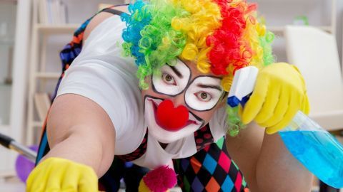 Ein Clown reinigt den Boden mit einem Schwamm und Reinigungsmittel, umgeben von einem hellen Raum voller Möbel.