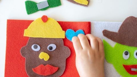 Ein Kind fügt bunte Filzgesichter in einem Filzbuch zusammen, umspielend mit kreativen Figuren auf einem Tisch.