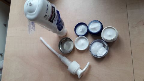 Eine Pumpspender-Lotion steht neben mehreren leeren Cremetöpfen auf einem Tisch, bereit für die Anwendung bis zum letzten Rest.