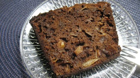 Ein Stück Kakao-Apfel-Mandel-Kuchen liegt auf einem klaren Teller und wird von einem Besteckstück umgeben.