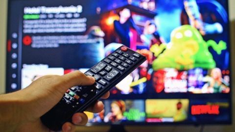 Ein Hand hält eine Fernbedienung, während ein Fernseher mit verschiedenen Serientiteln und Bildern von Charakteren im Hintergrund leuchtet.
