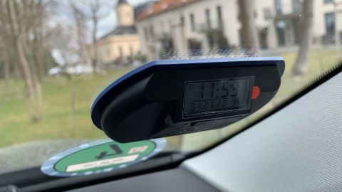 Eine elektronische Parkscheibe zeigt die Uhrzeit an und wird im Auto verwendet, um Parkzeiten zu regulieren, während im Hintergrund ein Gebäude sichtbar ist.