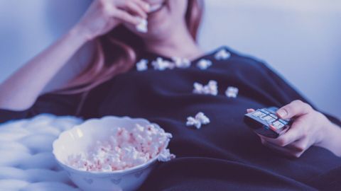 Eine Person sitzt entspannt auf einem Sofa, isst Popcorn aus einer Schüssel und hält eine Fernbedienung in der Hand.